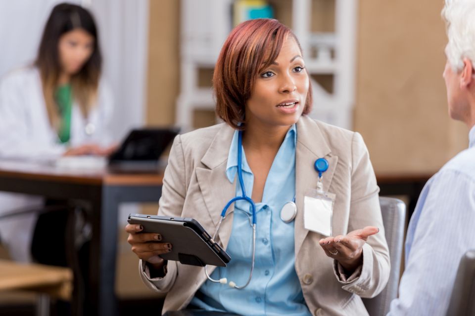 Pretty mid adult African American emergency medicine doctor gestures while discussing something with senior male colleague or patient. She gestures while holding a digital tablet. A female doctor is working in the background.