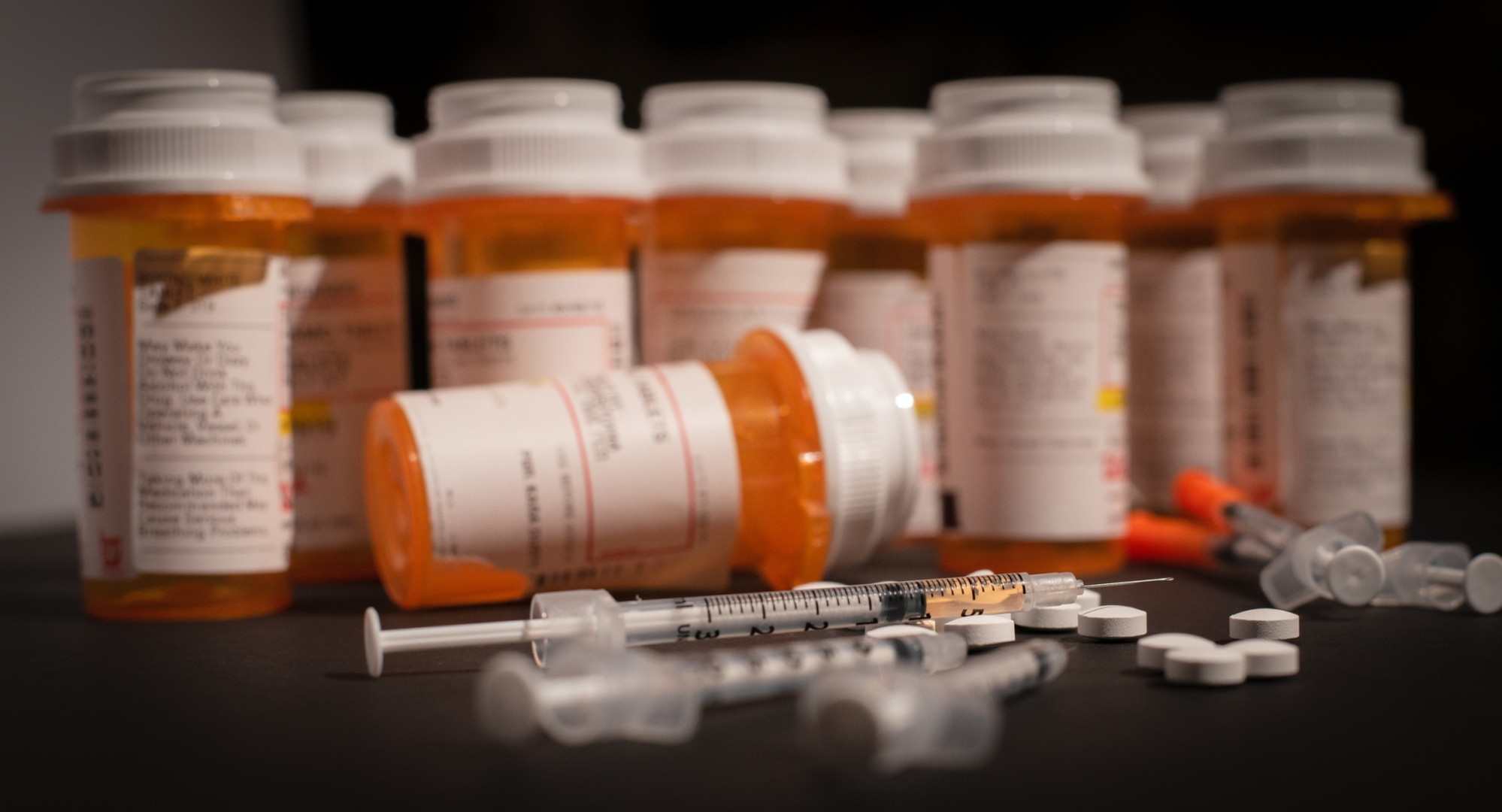 An injectable drug is loaded into a syringe while prescription medication is strewn about haphazardly.
