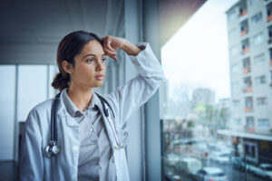 Shot of a young female doctor looking stressed out while standing at a window in a hospital