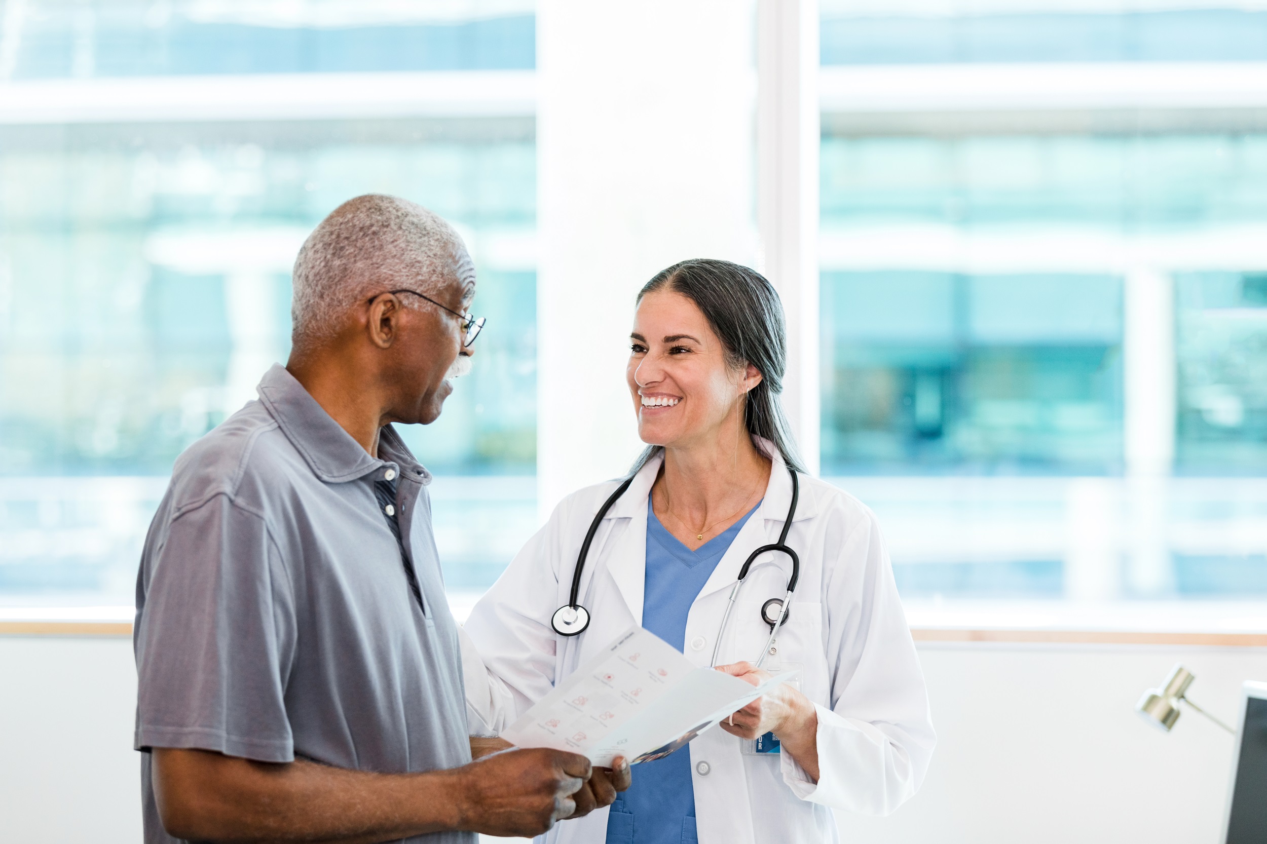 The mid adult female specialist smiles at her senior adult male patient as he asks a question about the information in the brochure.