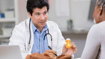 A mid adult male doctor sits at a table across from an unrecognizable patient.  He holds up a prescription bottle as he explains the benefits and risks of a new medication.