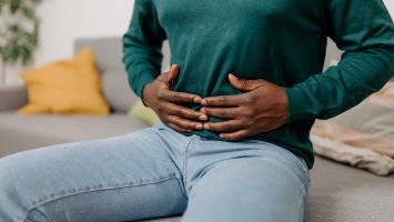 This image portrays a young African American man sitting on a sofa in his living room, holding his stomach due to digestive problems