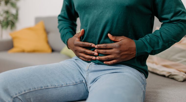 This image portrays a young African American man sitting on a sofa in his living room, holding his stomach due to digestive problems