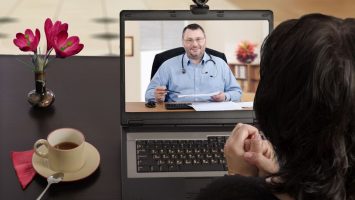 Blackhaired woman sits at a cafe table and consults telemedicine doctor by laptop computer with her back to camera. In monitor, male physician reviews medical laboratory results with her. Horizontal shot on indoors blurred background